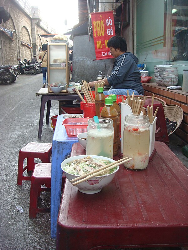 A pavement phở stall in Hanoi under a PHỞ BÒ GÀ sign, with low stools and jars of pickled garlic